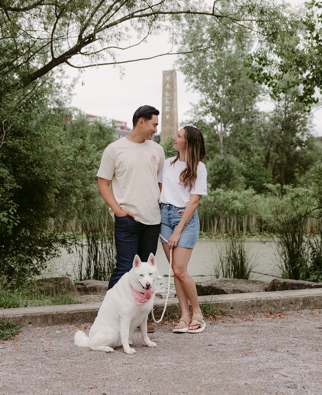 Couple standing together with their white dog wearing a pink bandana at Evergreen Brick Works in Toronto, smiling and enjoying a summer photo session by the pond.