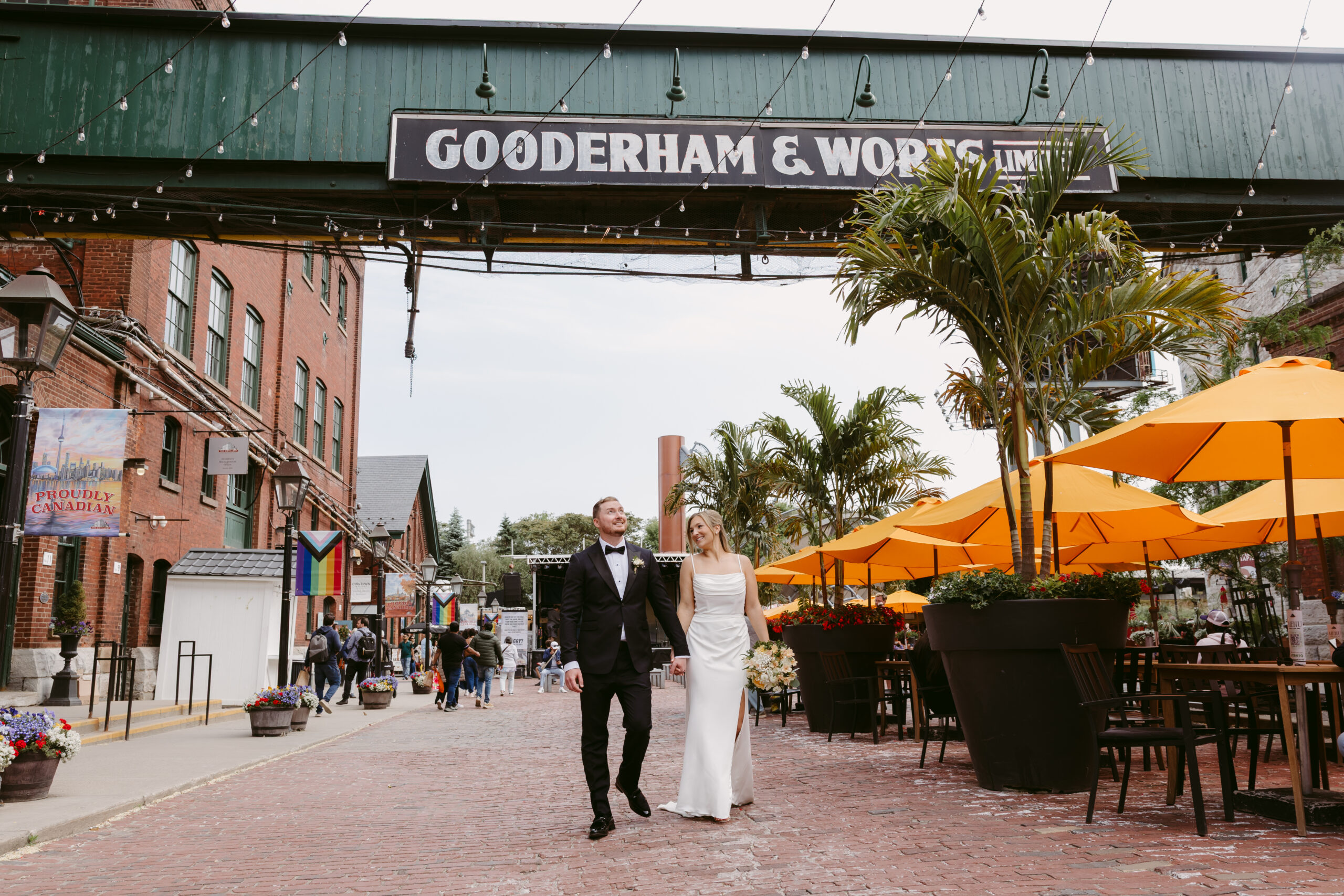 Bride and groom walk hand in hand through the Distillery District in Toronto, under the Gooderham & Worts sign, surrounded by vibrant umbrellas and brick buildings.