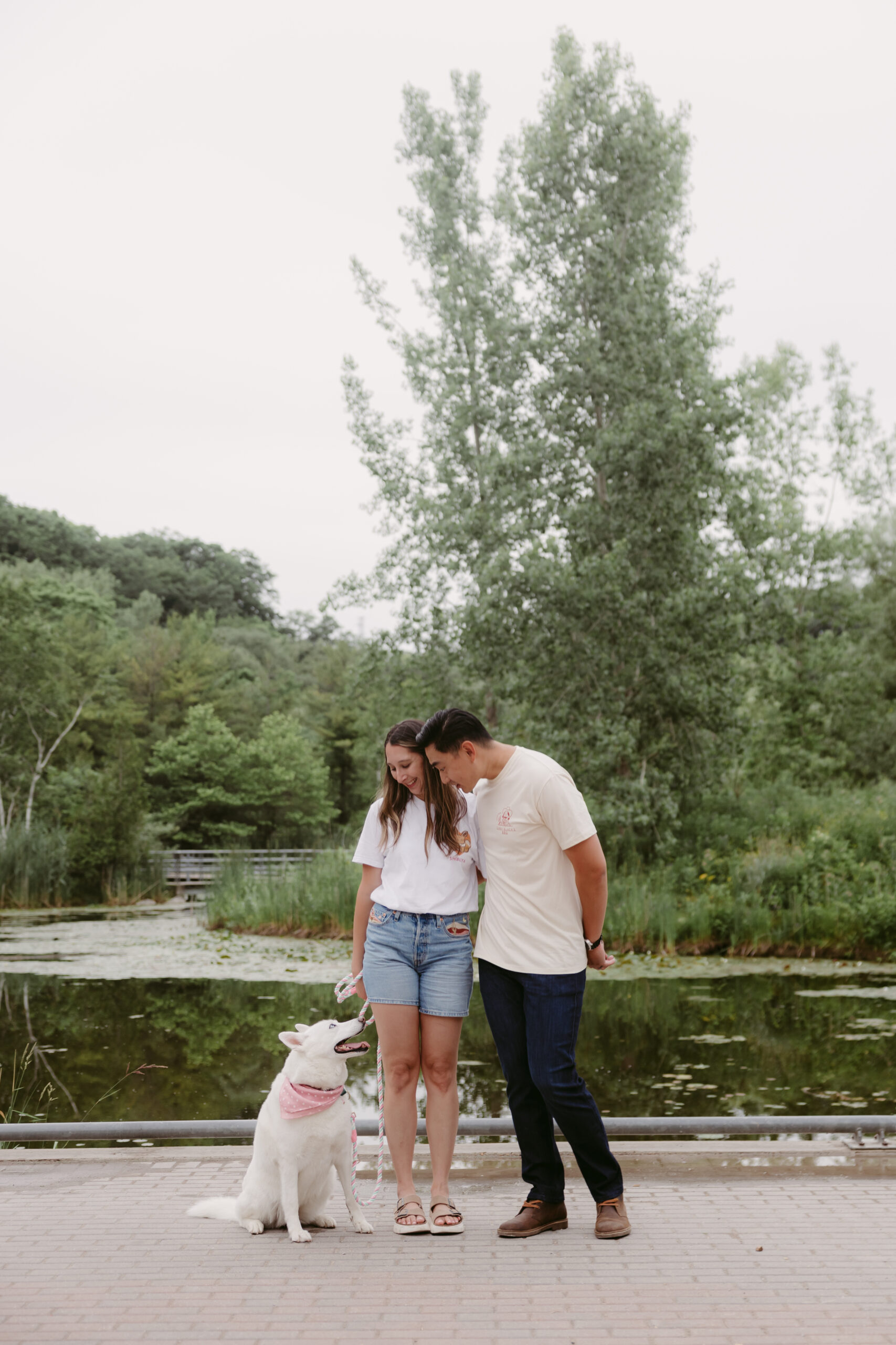 Couple standing together with their white dog wearing a pink bandana at Evergreen Brick Works in Toronto, smiling and enjoying a summer photo session by the pond.