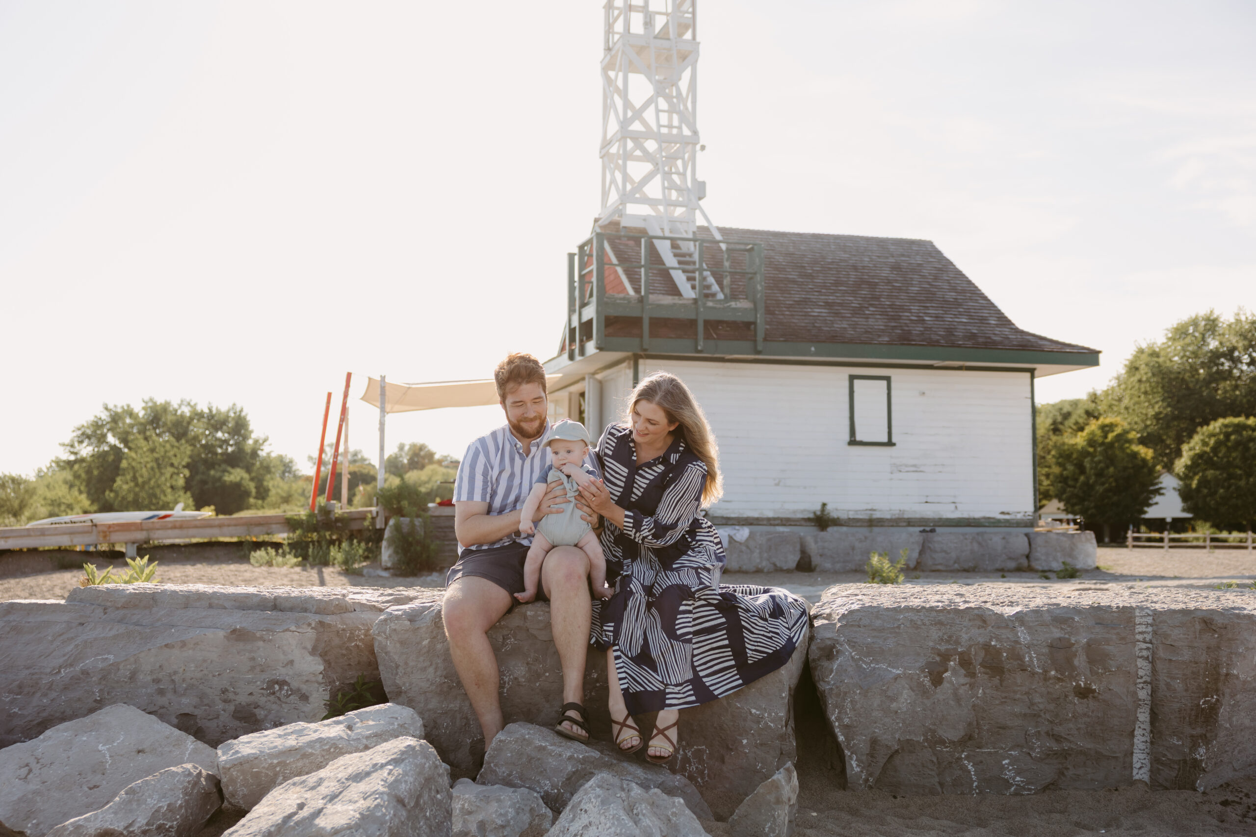 Family baby session at Toronto Beaches with parents sitting on rocks, holding their baby in front of the historic lifeguard station.