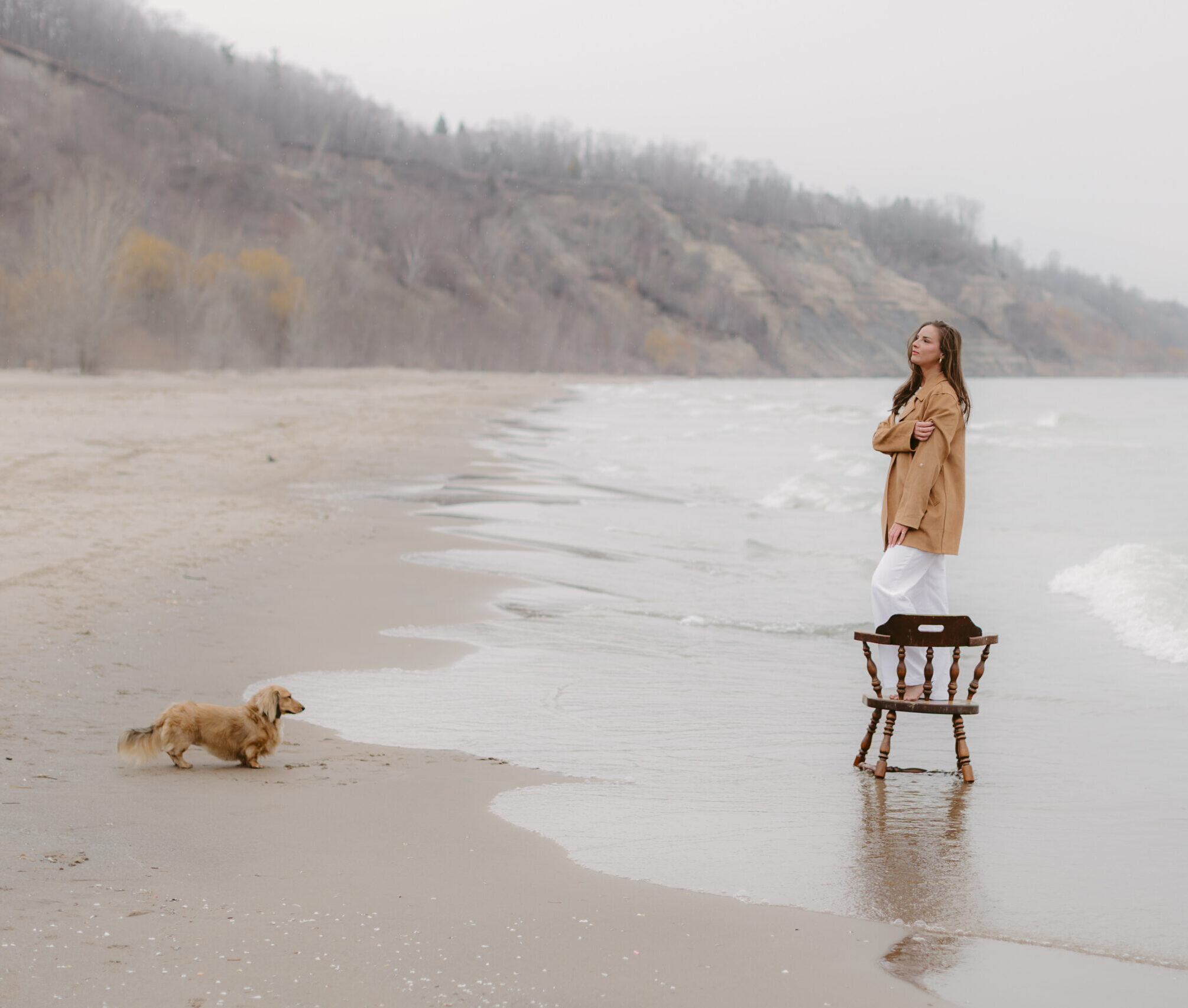Women standing on a wooden chair on the shoreline while her dog looks at her.