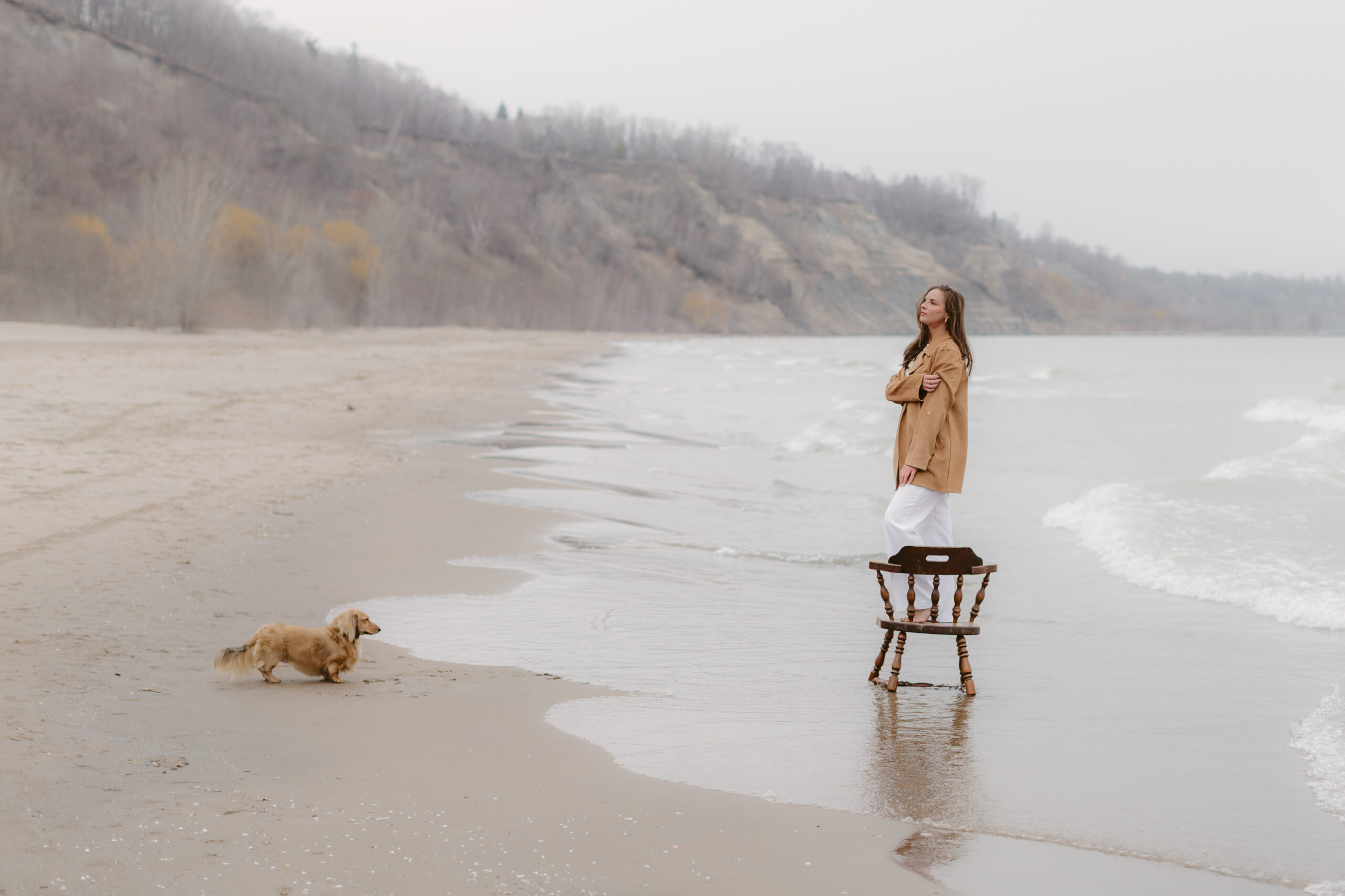 Women standing on a wooden chair on the shoreline while her dog looks at her.